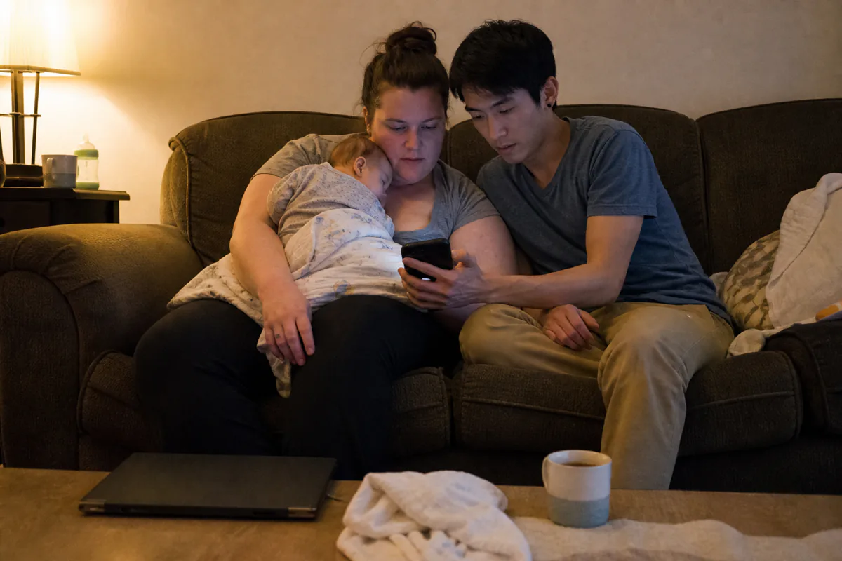 A couple on a sofa both looking at the same phone, a baby asleep on one of them
