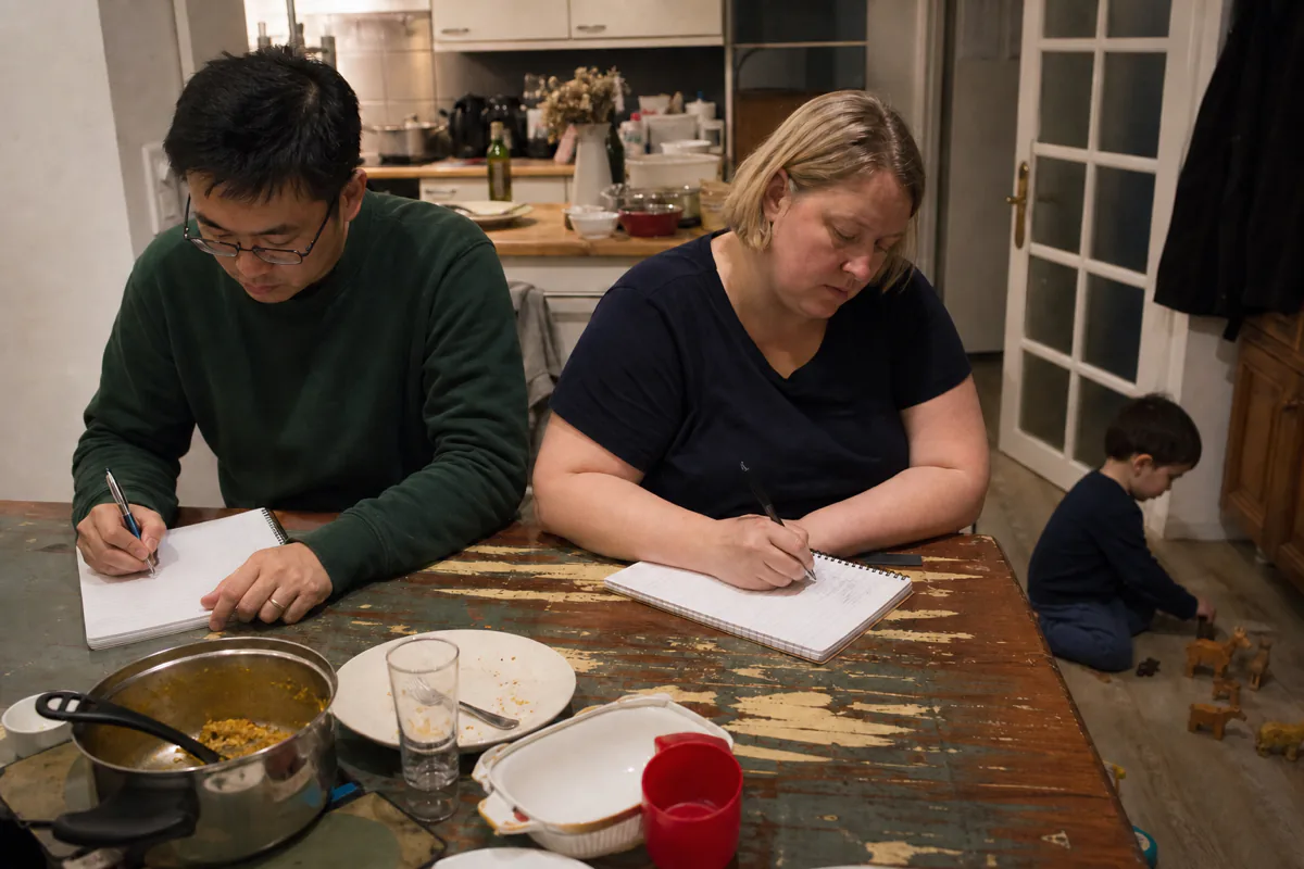 A couple at a kitchen table each writing in their own notebook, a toddler playing on the floor