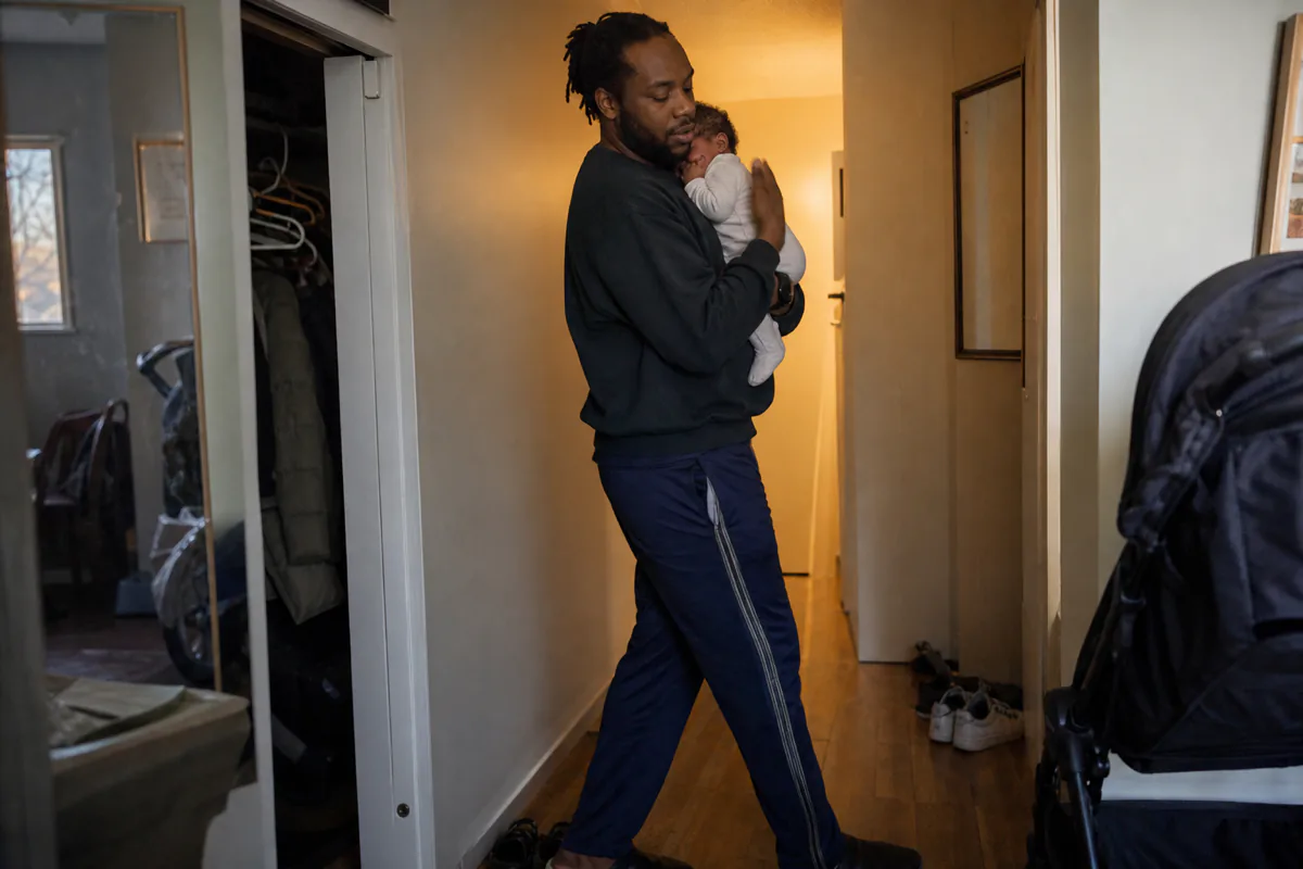 A parent walking a crying newborn along a hallway at dusk, patting their back