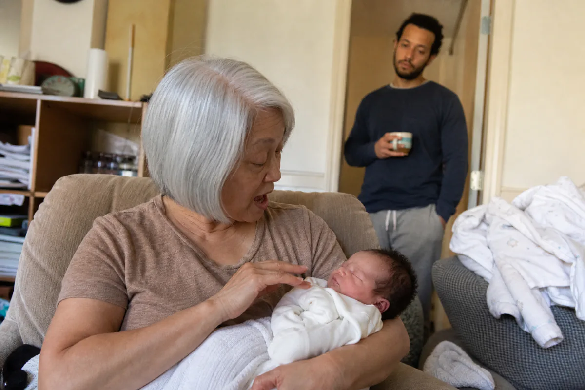 A grandparent in an armchair holding a newborn and talking, a parent watching from the doorway