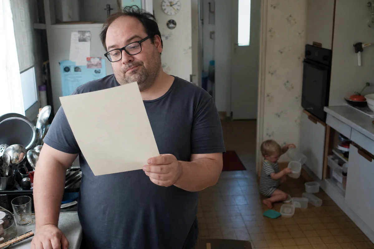 A parent in a kitchen reading a document while a toddler empties a cupboard behind them