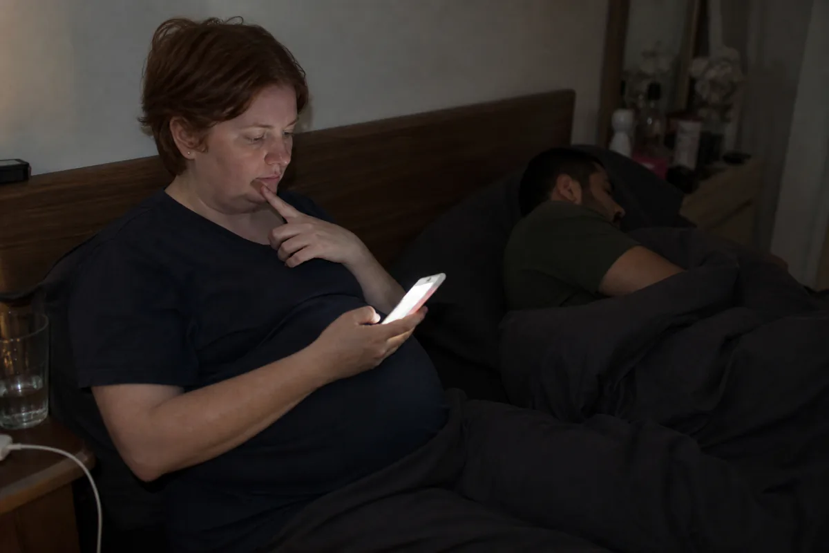 A pregnant woman awake at night, her face lit by a phone, her partner asleep behind her