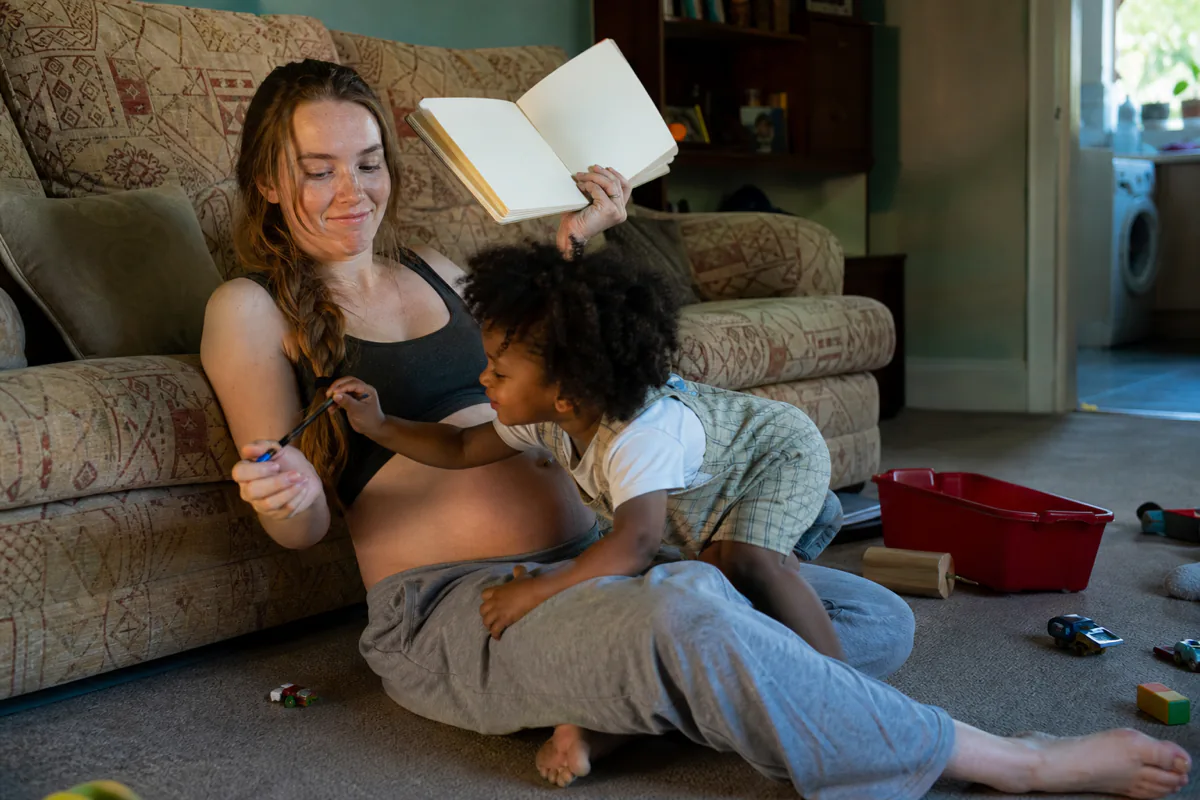 A pregnant woman with a notebook while a toddler climbs across her legs to reach the pen