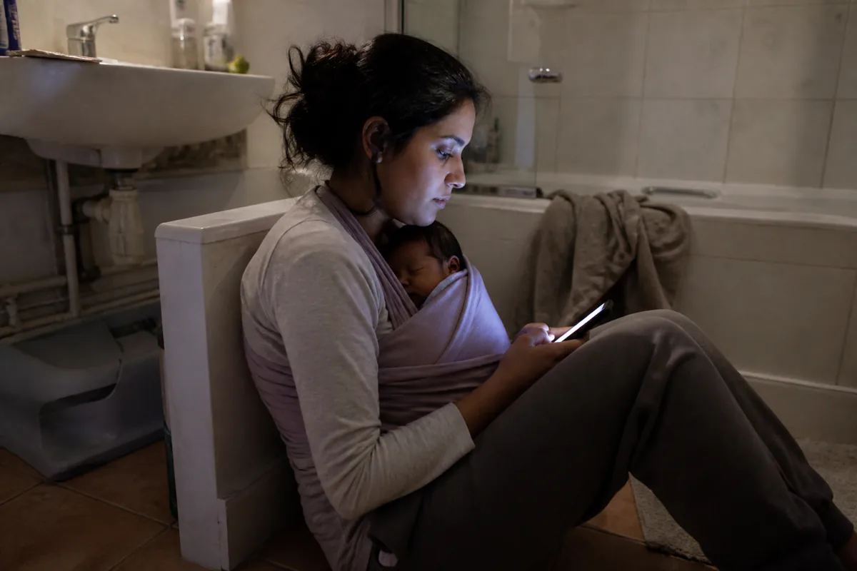 A parent on a bathroom floor at night holding a phone, a newborn asleep against their chest