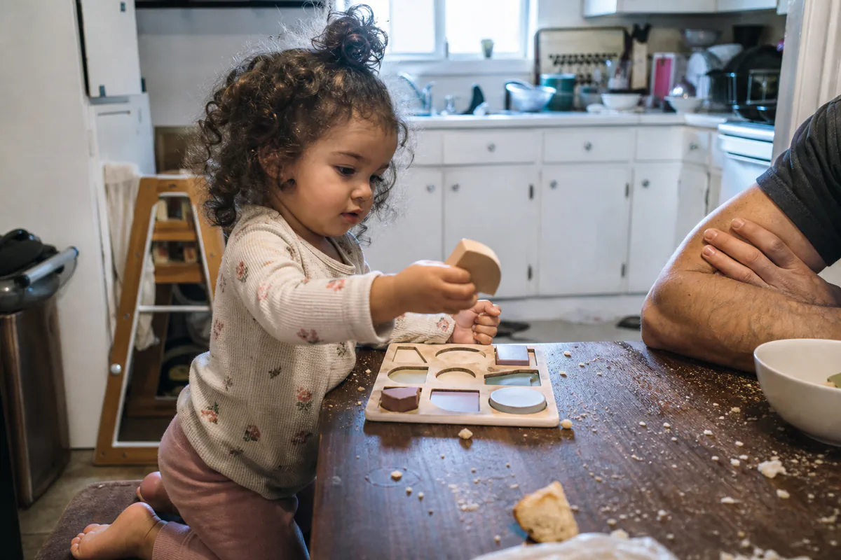 A toddler kneeling at a kitchen table pushing a wooden shape into a puzzle board